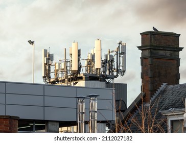 5g mobile phone radio mast transmitter at sunset in residential town centre with old 4g and 3g antennas also attached.  Buildings below with bright sky and city rooftops.