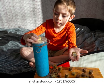 5-year-old Boy Puts 50 Euro Cent Coin In Selfmade Savings Jar,sit On Bed,wooden Box With Scattered Coins Columns In Front Of Him,sunny Day,hard Light.Learning Money,financial Literacy.Selective Focus.