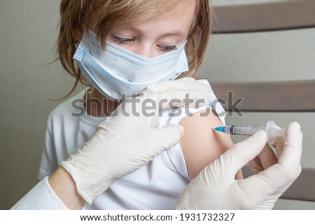 A 5-year-old Caucasian girl in a medical mask receives a vaccine, peeping with eyes down on the syringe