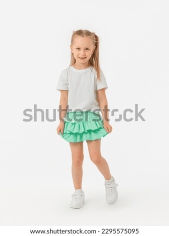 A 5-year-old girl with long hair looks expressively at the camera in a T-shirt and holds on to a skirt-shorts, bending her knee in sneakers on a white background. photo