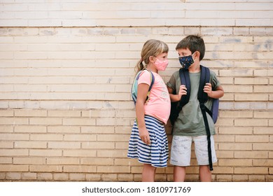 6 Year Old Caucasian Boy And Girl Wearing Colorful Protective Masks And Backpacks Talking With A Wall In The Background. Preschoolers On The First Day Of School After The Coronavirus Pandemic.