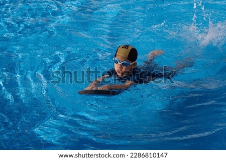 6 years old young boy who a swimmer wearing goggle with dark blue suit and yellow striped black cap. Lonely child warm up with swimming kickboard before practicing to swim in the blue swimming pools.
