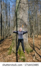 60 Year Old Sporty Woman Hugging Old Mighty Tree In The Forest In Ukraine On A Sunny Day. Back View. The Symbiosis Of Man And Nature.