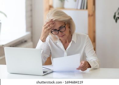60 years old serious businesswoman reading document holding ache at work. Tired woman employee looking at report on paper near modern laptop having headache. Stressful business deal concept. - Shutterstock ID 1694684119