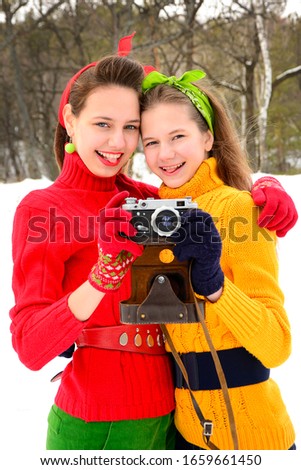 60 years style. Two girls in red and yellow sweaters playfully smile at the lens and are holding an old camera. Vertical