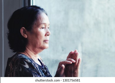 60S Or 70s  Years Old Asian Elderly Woman Sad By The Window.Granny Touch The Window Glass And Looking Outside,she May Worried Or Thinking Of Something Or Waiting For Someone.Concept Of Lonely Elderly.