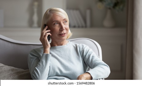 60s Blonde Woman Sit On Couch Holds Phone Chatting With Friend Having Pleasant Distant Conversation Talking With Grown Up Children, Elder Generation And Modern Technologies Everyday Easy Usage Concept