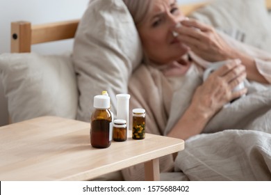 60s elderly woman lying in bed holding cup blow runny nose using handkerchief close up focus on bedside table full of antiviral medications, bottle of pills, grippe influenza, caught a cold concept - Powered by Shutterstock