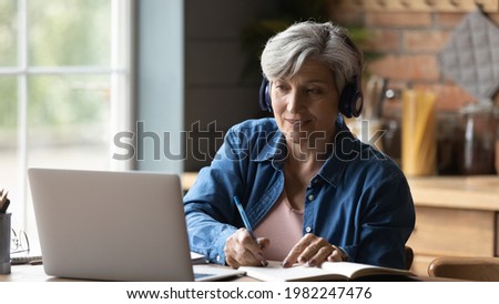 60s woman sit at table wear headphones take notes gain new skills use laptop and internet resources, on-line services. Video call event, modern tech, counselling, receive information remotely concept