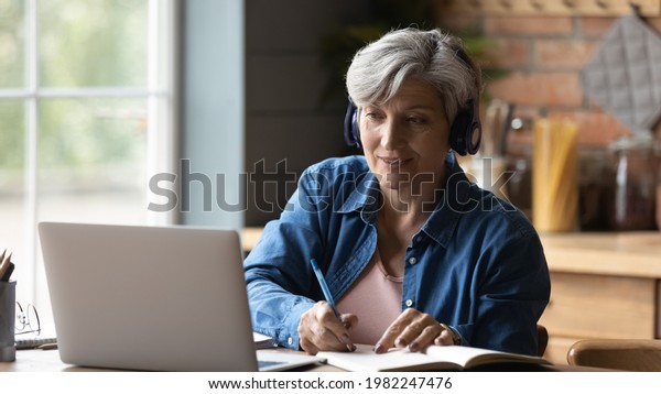 60s woman sit at table wear headphones take notes\
gain new skills use laptop and internet resources, on-line\
services. Video call event, modern tech, counselling, receive\
information remotely\
concept