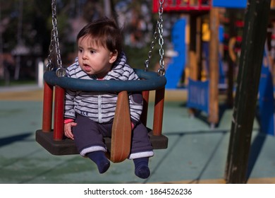 
6-month-old Baby, Sitting On A Swing, Swinging Happily And Happily. They Are In A Playground, Wearing A Navy Blue Striped Hoodie.