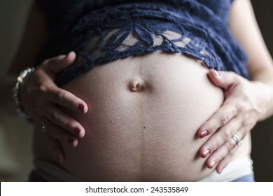 A 7 1/2 months pregnant woman wearing jeans and a blue lace shirt sits, holding her hands on her bare belly.
