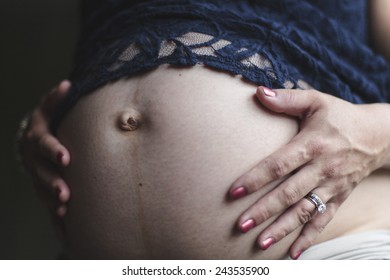 A 7 1/2 months pregnant woman wearing jeans and a blue lace shirt, holding her hands on her bare belly.