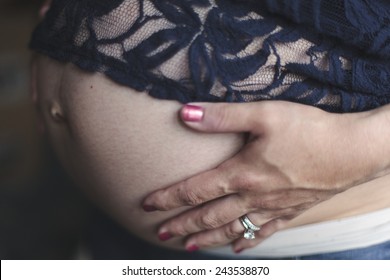 A 7 1/2 months pregnant woman wearing jeans and a blue lace shirt stands in front of a window holding her hands on her bare belly.