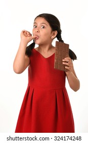 7 Or 8 Years Old Hispanic Girl Wearing Red Dress Holding Big Chocolate Bar Eating In Happy And Excited Face Expression Licking Her Fingers In Sugary Nutrition And Kids Loving Sweet Concept