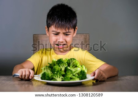 7 or 8 years old upset and disgusted Asian kid sitting on table in front of broccoli plate looking unhappy rejecting the fresh food in child hate green vegetables concept and healthy nutrition 
