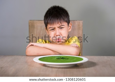 7 or 8 years old upset and disgusted Asian kid sitting on table refusing to eat spinach thick soup looking unhappy rejecting vegetarian food in child hate green vegetables and healthy nutrition 