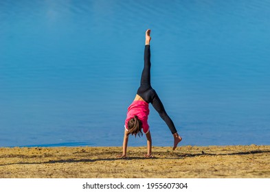 7 years old girl with pink shirt and sportswear doing a cartwheel at the beach during spring with blue water as background.
