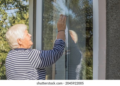 70 Year Old Woman Cleans Windows From Stains Using Rag And Spray Cleaner. Caucasian Elderly Woman Is Cleaning House, Doing Household Chores.