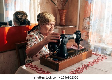 70 years old woman sewing at home, senior seamstress sitting at desk in front of sewing machine, her hands pushing pink fabric