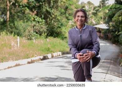 75-year-old woman in sportswear doing knee stretches outdoors, smiling and looking at the camera while staying active and healthy.