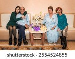 76-year-old man and 71-year-old woman celebrate their golden wedding anniversary, sitting on a sofa in the registry office, embraced by their daughter and granddaughter, holding a bouquet and medal