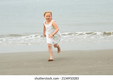 a 7-year-old girl in a white dress plays on the seashore collecting shells and running from the waves  - Shutterstock ID 2134910283