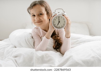 8 years girl holding alarm clock 7 am sitting on bed under white blanket, sleep at morning. Time go to school, wake up early at home - Powered by Shutterstock