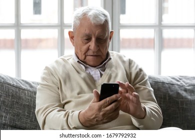 80s grey haired old man sit on sofa hold cellphone looking at screen makes call to physician, writing or sending sms to friend. Modern wireless technology easy usage of older generation people concept