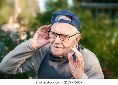 An 88-year-old Man In A Baseball Cap And Glasses Listens. Portrait Of An Old Farmer With Poor Hearing.