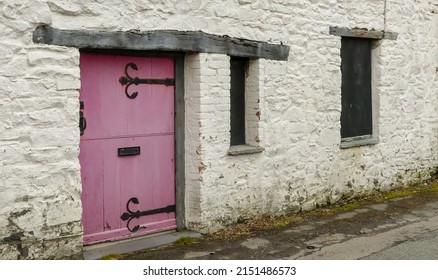 Abandoned, boarded up frontage of an old cottage. Original weathered oak lintels above windows and door. Pink painted stabledoor entrance with old ironwork furniture. Whitewashed exterior with moss.