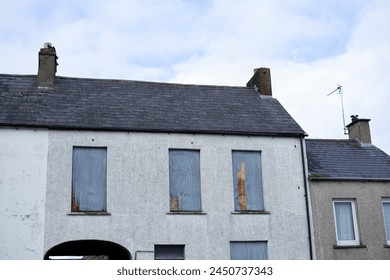 Abandoned boarded up house home building windows wood plywood arched walkway driveway roof shingles antenna chimneys clouds in blue bright sunny sky Irish small town city near Belfast sleepy  - Powered by Shutterstock