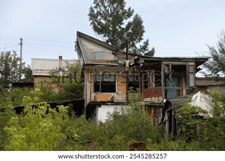Abandoned dacha with a unique, improvised structure featuring mismatched materials and colorful panels. Surrounded by overgrown greenery, it has a quirky, forgotten appearance.