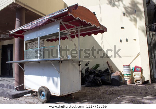 Abandoned food cart\
chained to an abandoned building. To the side, black trash bags and\
empty containers.