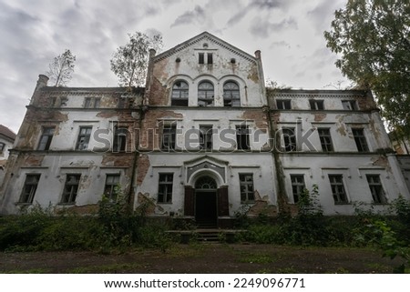 Abandoned, leaning building of an old psychiatric hospital. The cracked plaster on the walls exposes the red brick. Windows are broken, glass sticks out of the openings. Gloomy sky in the background