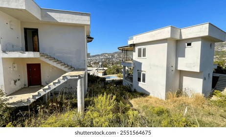 Abandoned modern-style houses surrounded by overgrown vegetation under a clear blue sky, concept of urban decay and redevelopment, suitable for real estate, architecture, and environmental campaigns. - Powered by Shutterstock