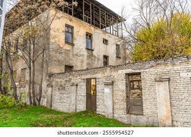 An abandoned multi-story building with weathered walls and broken windows, surrounded by leafless trees. A dilapidated brick wall with rusty doors in the foreground contrasts with the vibrant green - Powered by Shutterstock