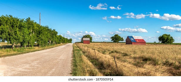 An abandoned old barn in Rural Texas with the state flag painted on its roof sits in a farmland community framed by fields and a dirt road.