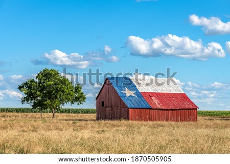 An abandoned old barn with the symbol of Texas painted on the roof sits in a rural area of the state, framed by farmland.

