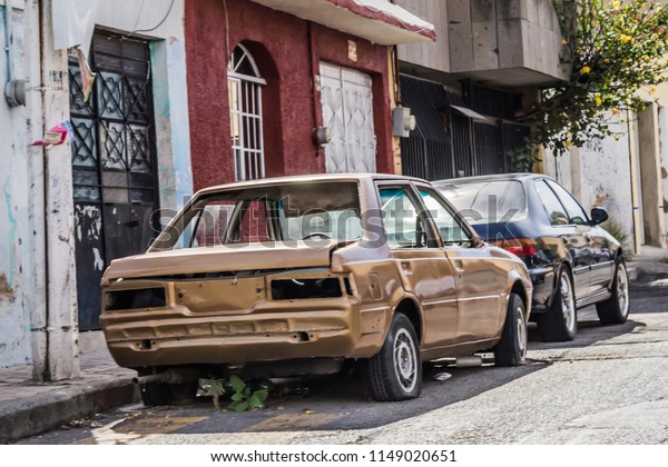 Abandoned old car without glass, with\
broken taillights, flat tires and plants underneath, indicating how\
long it has been there, on some street in\
Mexico