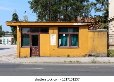Abandoned old dilapidated yellow security guard entrance to workplace building and cracked wooden doors   facade in front asphalt road and large trees   construction material in background