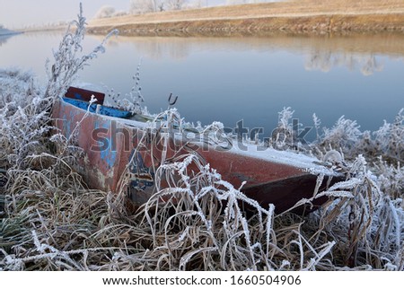 Abandoned old small boat on the river shore in winter. A rusty rowing boat full of dirt and water. Winter cold morning, hoarfrost on boat and grass.