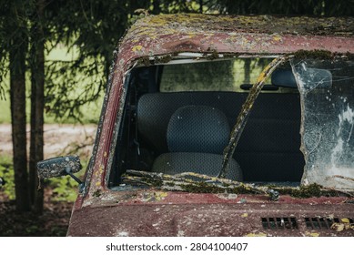 Abandoned red car with a broken windshield and moss-covered exterior, showcasing an empty interior, set in a rural area with trees in the background