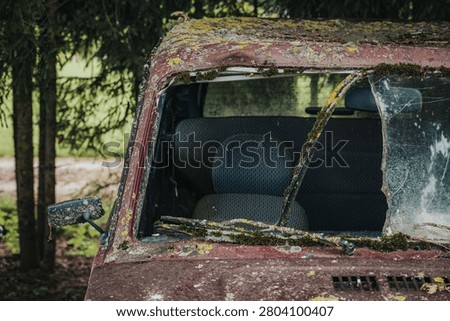 Abandoned red car with a broken windshield and moss-covered exterior, showcasing an empty interior, set in a rural area with trees in the background