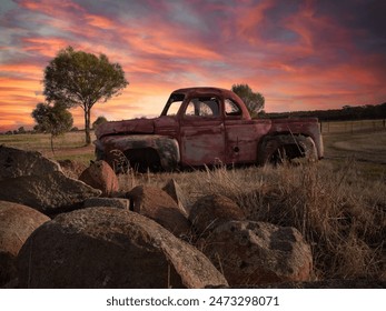 A abandoned red and rusty American truck pickup sitting in a farm field with a red and gold sunset in the background , rocks are in the foreground with a couple of small trees framing the vehicle  - Powered by Shutterstock