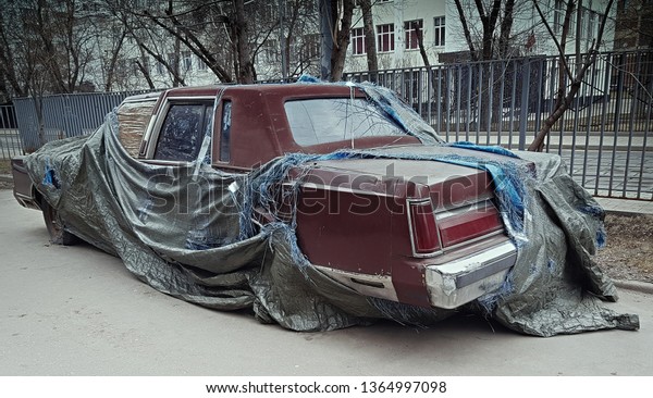Abandoned rotting car under a torn canvas cover on\
the street