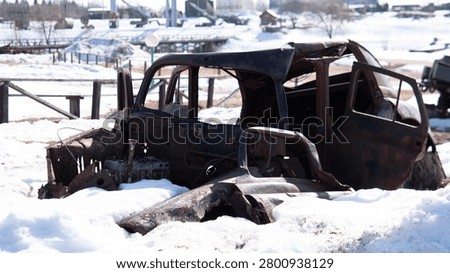 Abandoned rusting car shell with missing doors and collapsed roof half buried in melting snow beside rural wooden fence; front seats and dashboard exposed