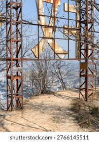 An abandoned, rusting metal framework with large peeling letters stands over a sandy dirt trail on a hilltop overlooking a distant urban landscape. The scene evokes a sense of decay, forgotten landmarks, and urban exploration.