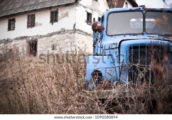An abandoned, rusty lorry with a rundown\
house in background.