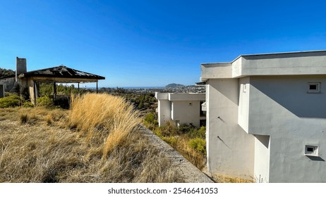 Abandoned structures with overgrown vegetation on a hillside overlooking a scenic coastal city under a clear blue sky, concept of urban decay, redevelopment, and environmental renewal projects. - Powered by Shutterstock